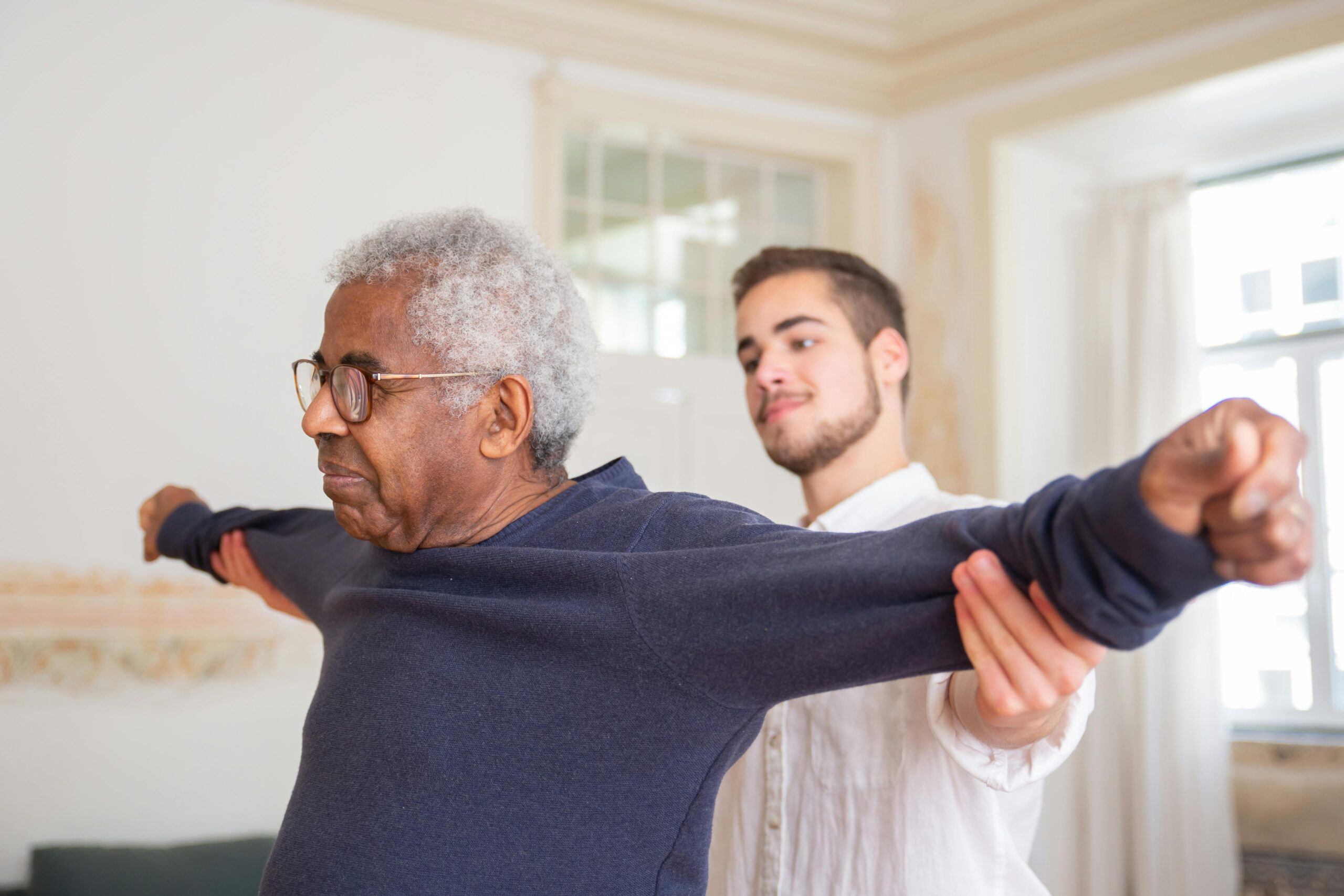 Find Physiotherapist Near You A senior man is assisted by a caregiver for stretching exercises indoors.